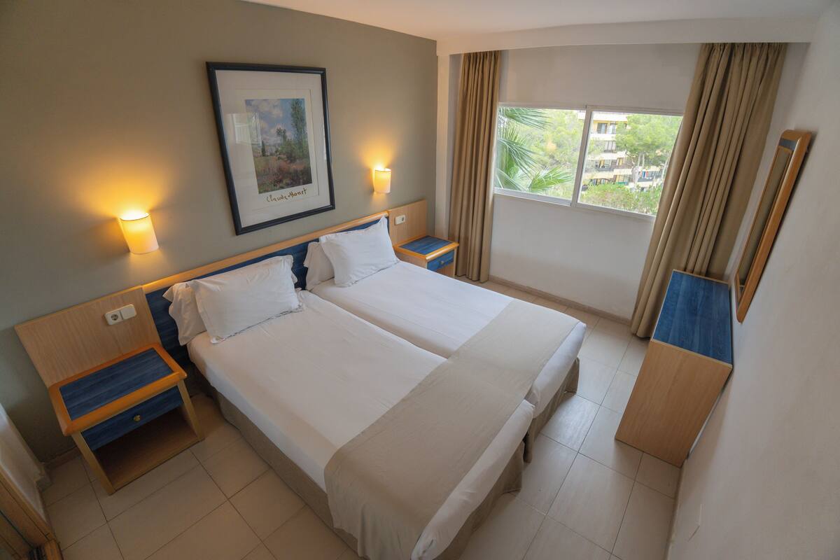 Ein Hotelzimmer mit zwei Betten, einem Nachttisch, einem gerahmten Bild, einem Fenster mit Vorhängen und einem Spiegel.