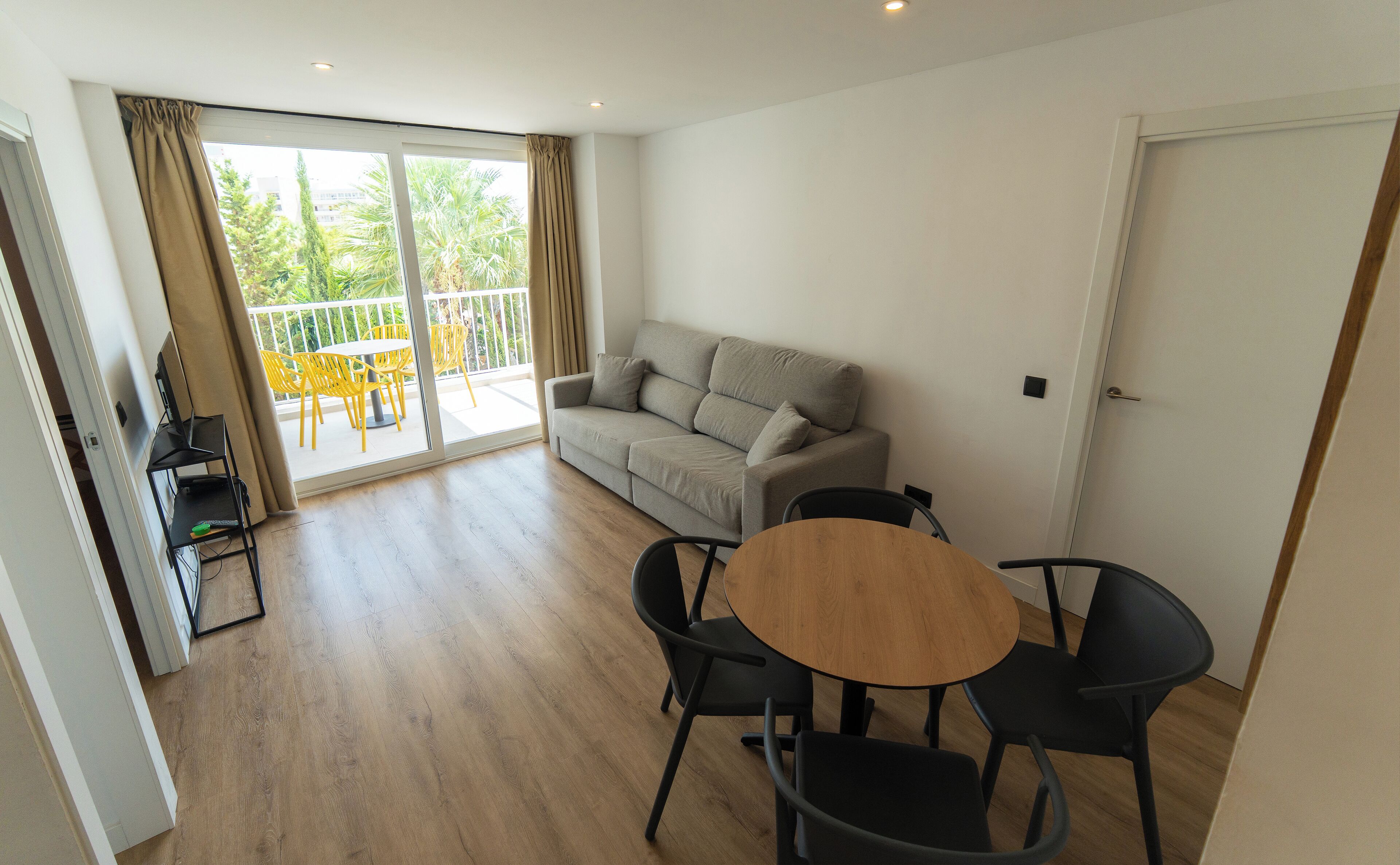 Un salon moderne avec un canapé gris, une table à manger en bois et des chaises noires. Il y a un balcon avec des chaises jaunes et un téléviseur posé sur un meuble.