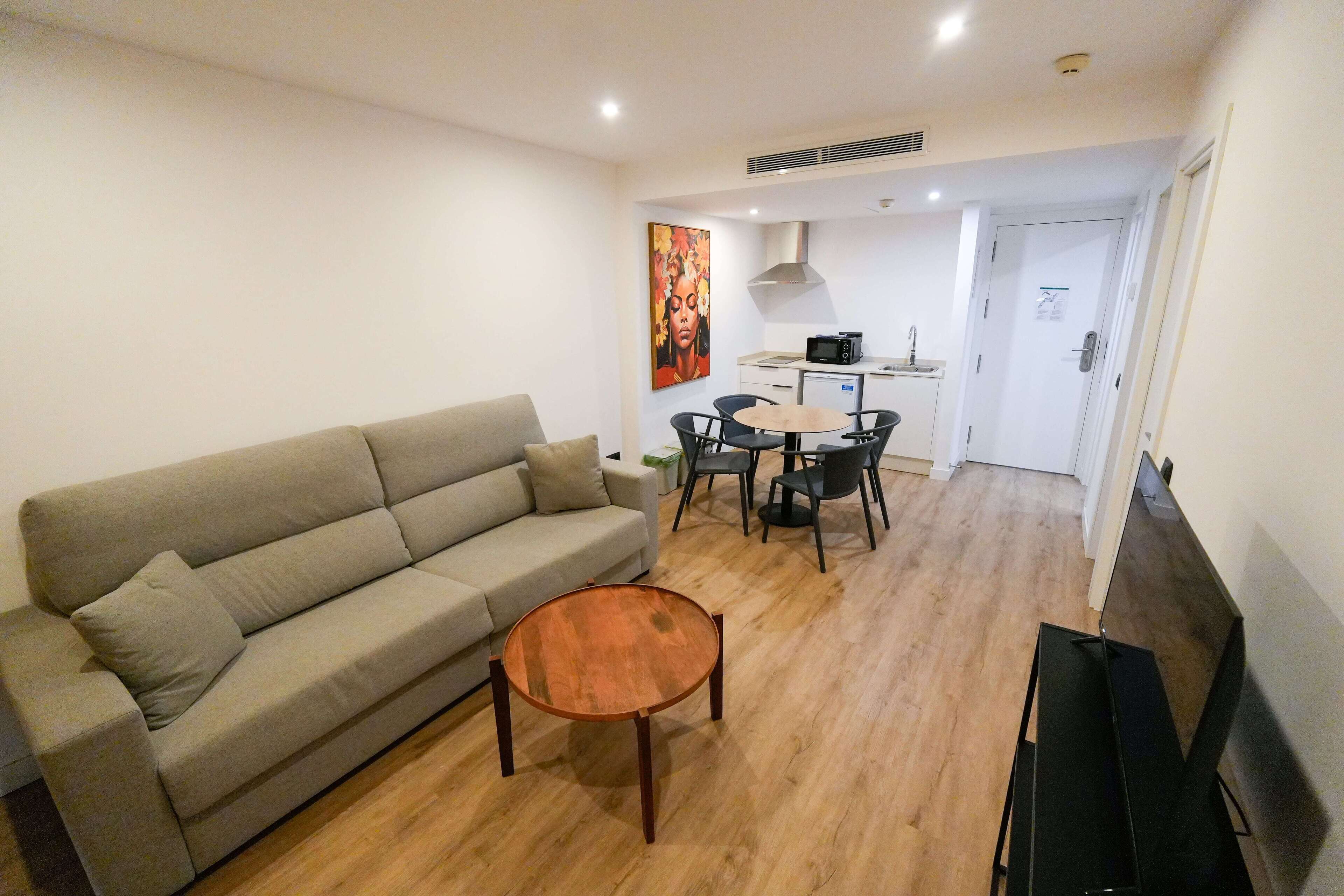Una sala de estar con un sofá, una mesa de centro y una zona de comedor con sillas y una pequeña mesa. Se observa una cocina al fondo con un microondas y un fregadero.