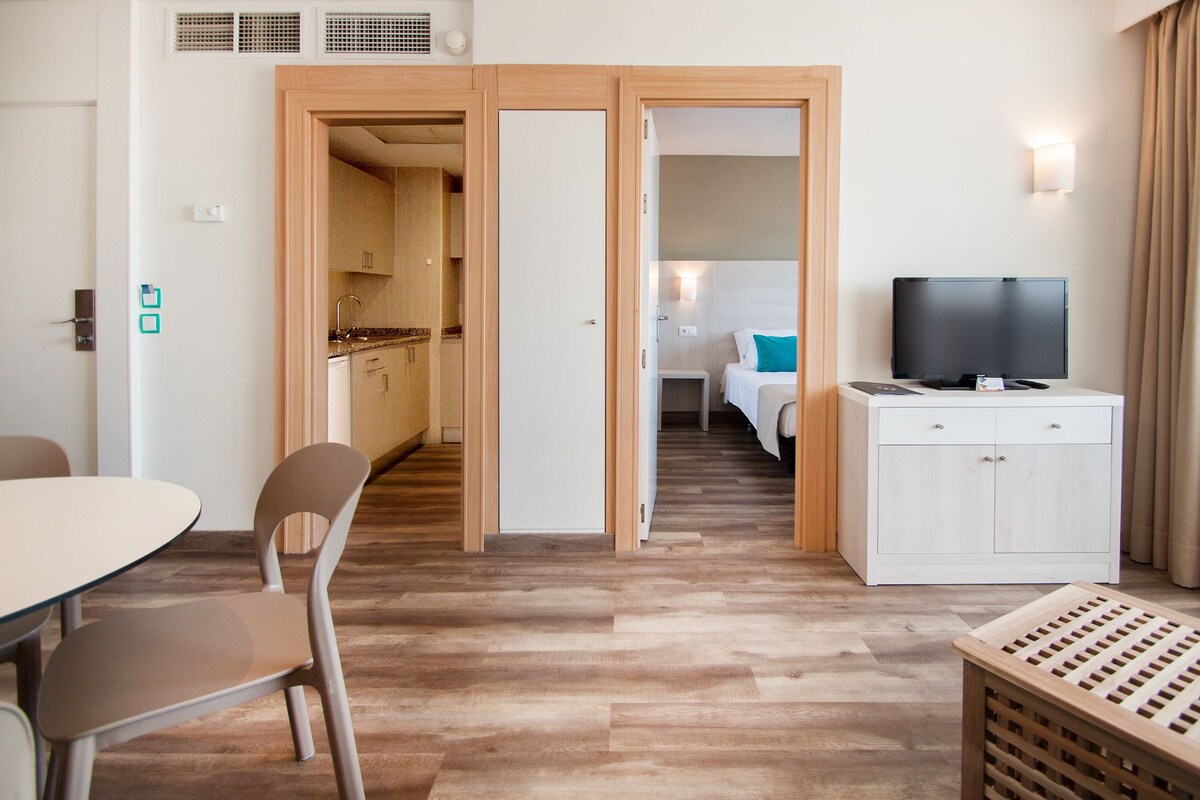 A hotel room with a wooden floor, a dining table with chairs, a TV on a cabinet, and a bed with a blue pillow.