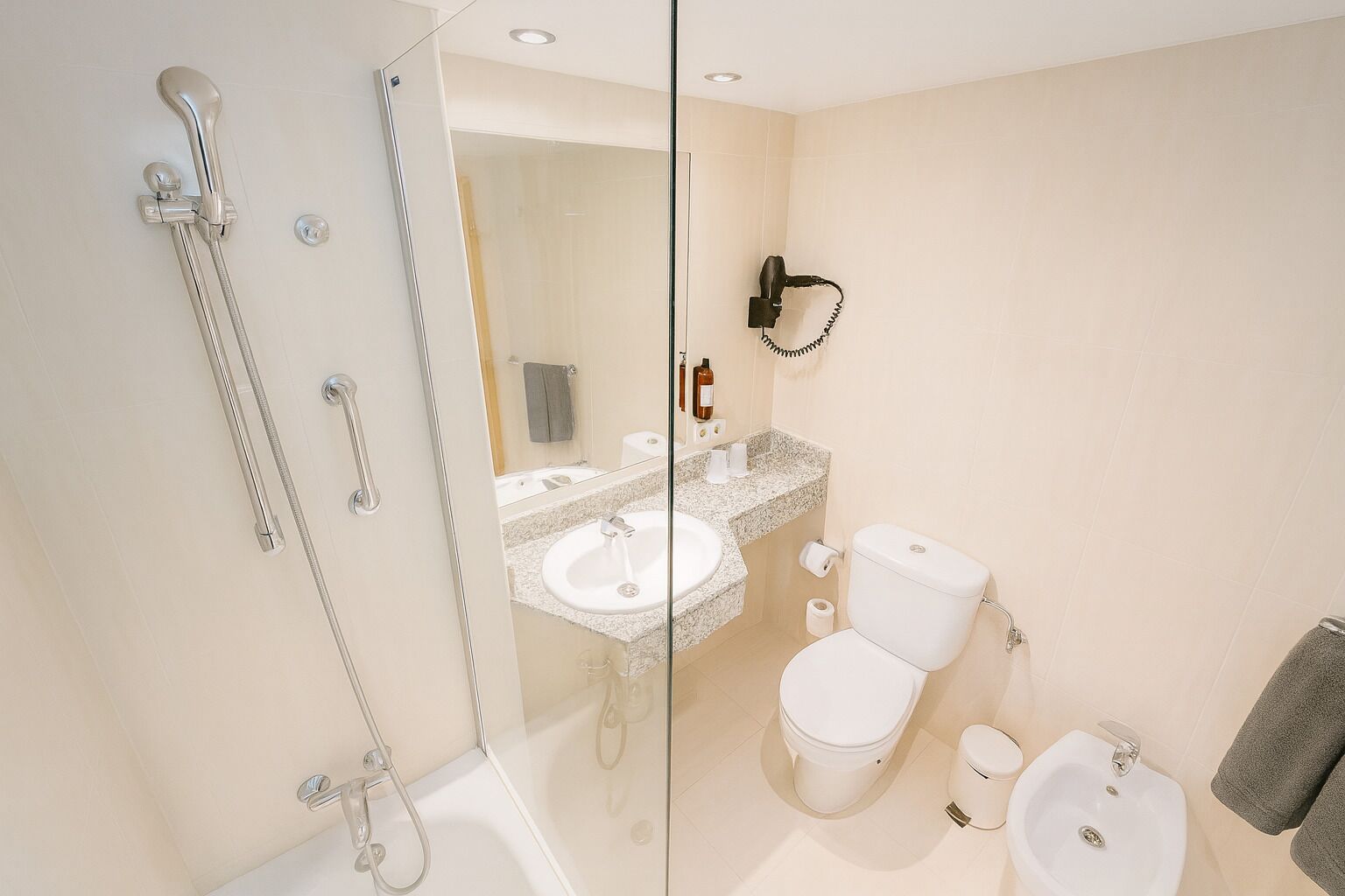 A modern bathroom with a glass-enclosed shower, a sink, toilet, bidet, and a hairdryer mounted on the wall.
