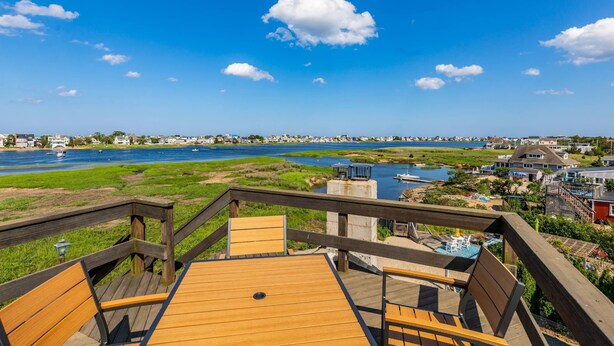 A wooden deck with a table and chairs overlooking a river, houses, and a clear blue sky.