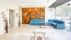 Een moderne lobby met een blauwe bank, een houten wand met geometrische patronen, en twee vazen met gedroogde bloemen.