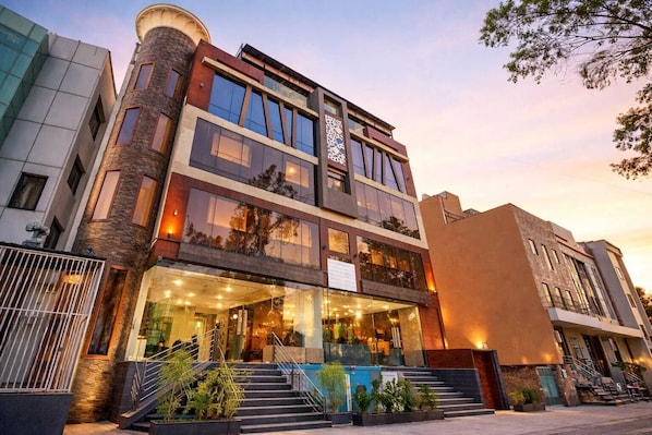 Un hotel de varios pisos con una entrada destacada, una escalera que conduce a la entrada y un área de piscina visible en primer plano.