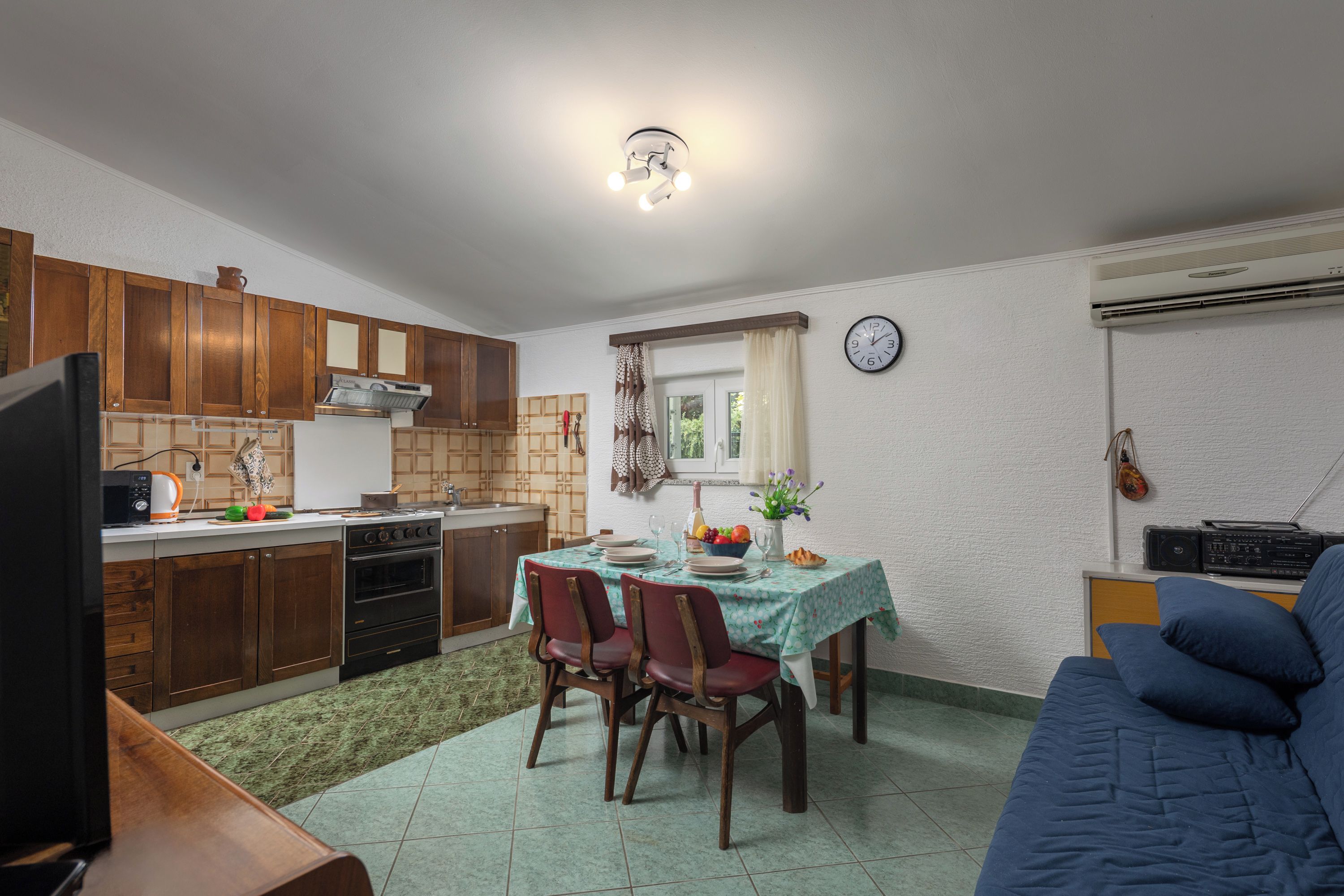 A compact kitchen with wooden cabinets, a dining table with chairs, a sofa, and a television.