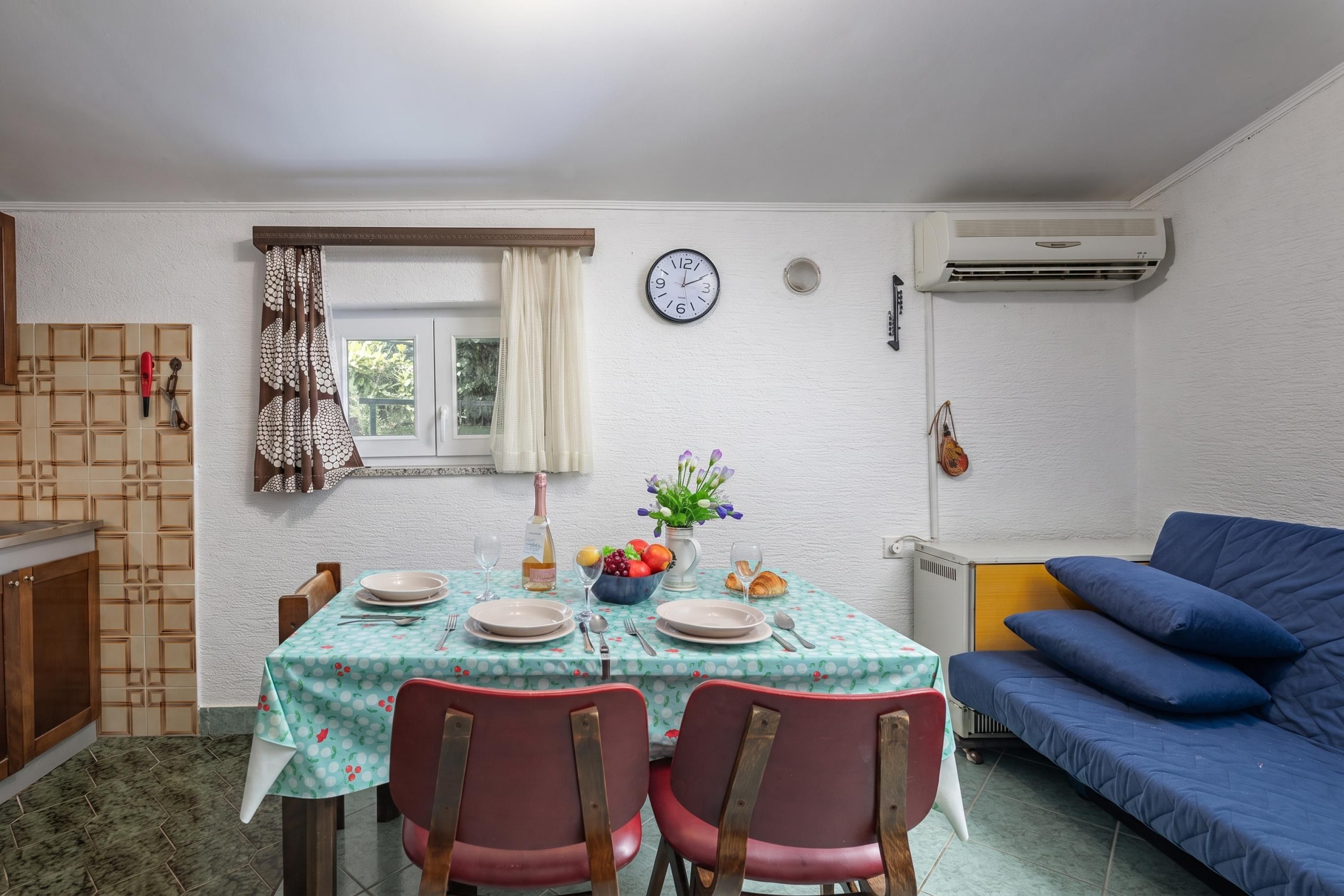 A dining area with a table set for two, a clock on the wall, and a blue sofa in the background.