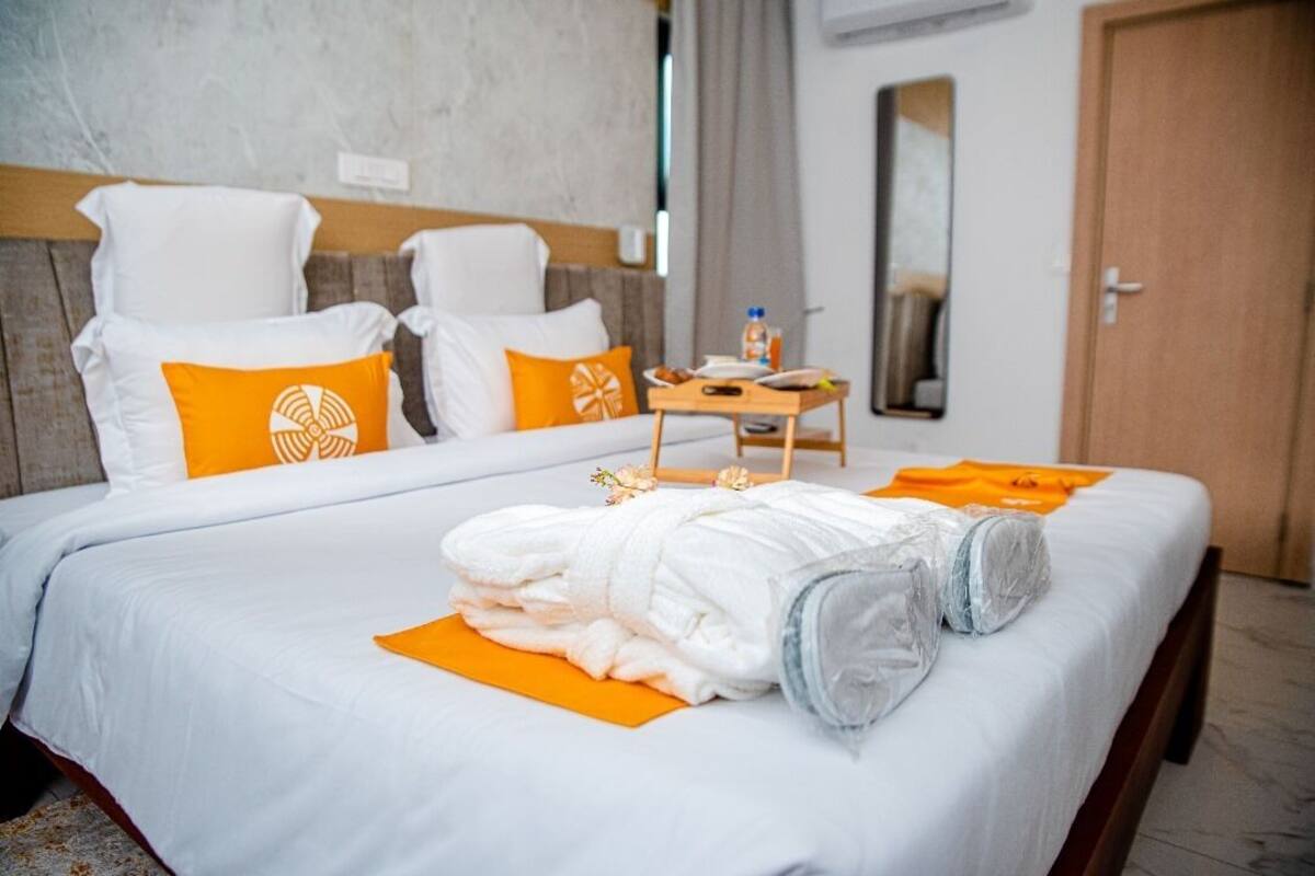 A hotel room with a bed, two white chairs, an orange mat, a wooden table with a bottle, and a mirror on the wall.