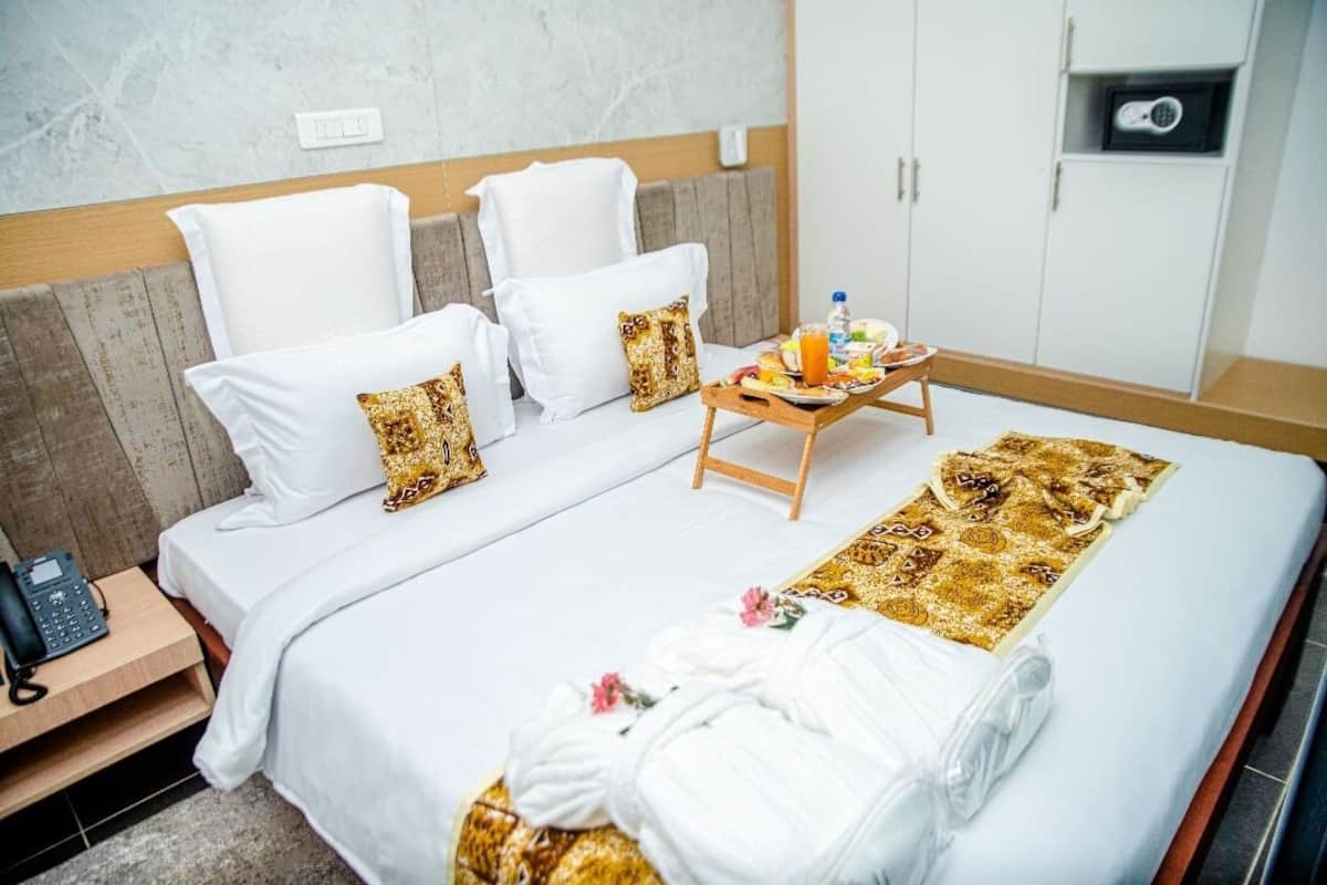 A hotel room with a bed, white bedding, a wooden headboard, a bedside table with a phone, a tray with breakfast items, and a towel on the bed.