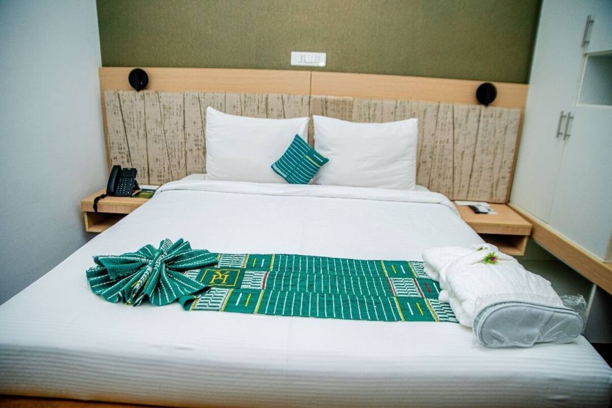 A neatly made bed with a green and white patterned blanket, white pillows, and a folded towel on the side.