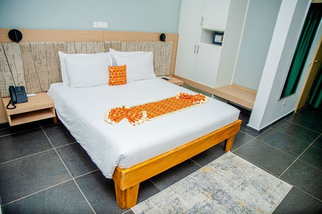 A neatly made bed with a white and orange patterned blanket, a wooden headboard, and a bedside table with a phone and a lamp.