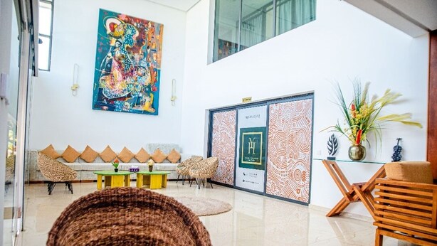 A spacious lobby with a high ceiling, a large colorful mural, a seating area with cushions, a glass display case, and a decorative plant. - HOTEL YATCHENIN YAMOUSSOKRO (Yamoussoukro)