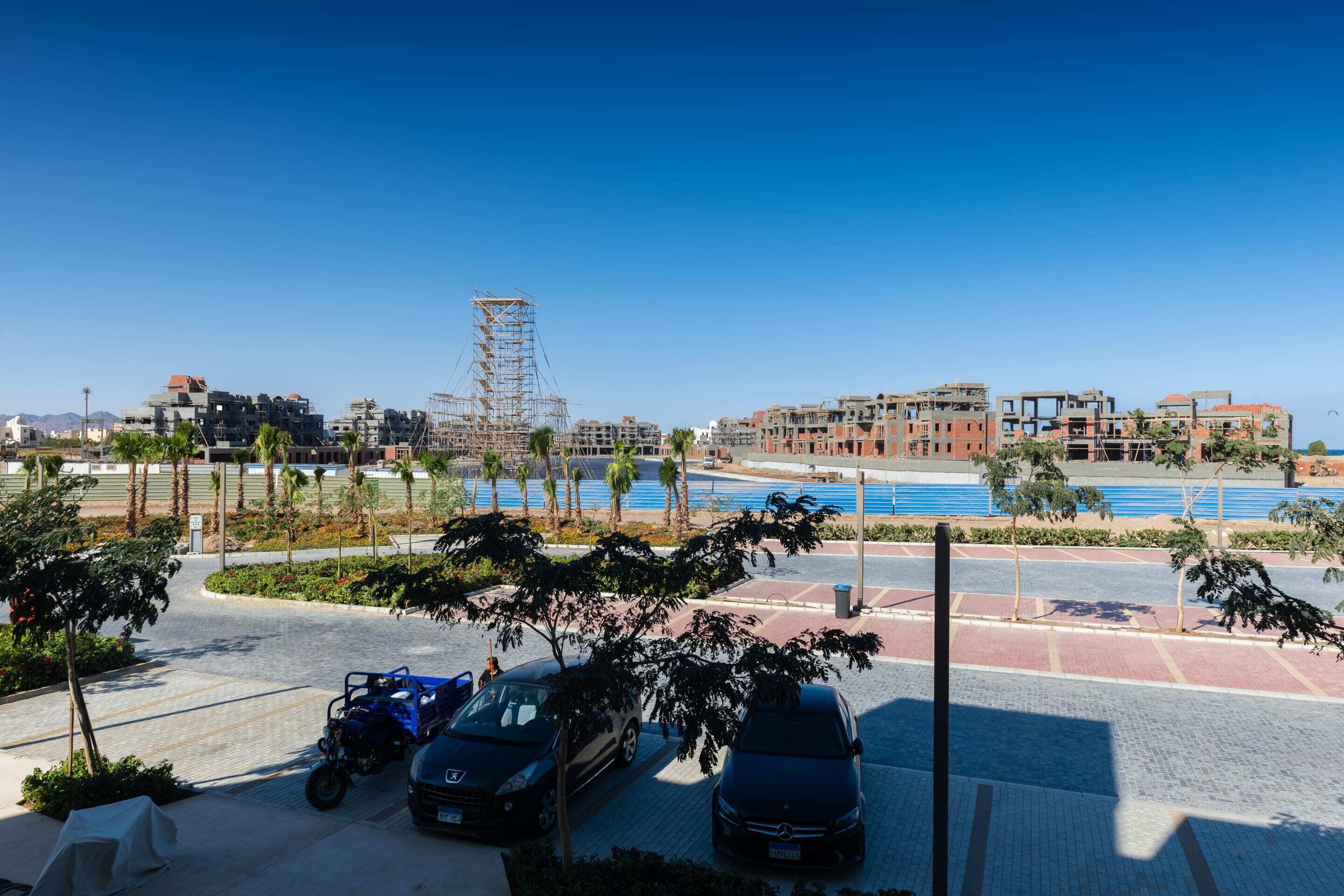 A parking area with several cars, a blue scooter, and a motorcycle. There are palm trees and a clear blue sky.