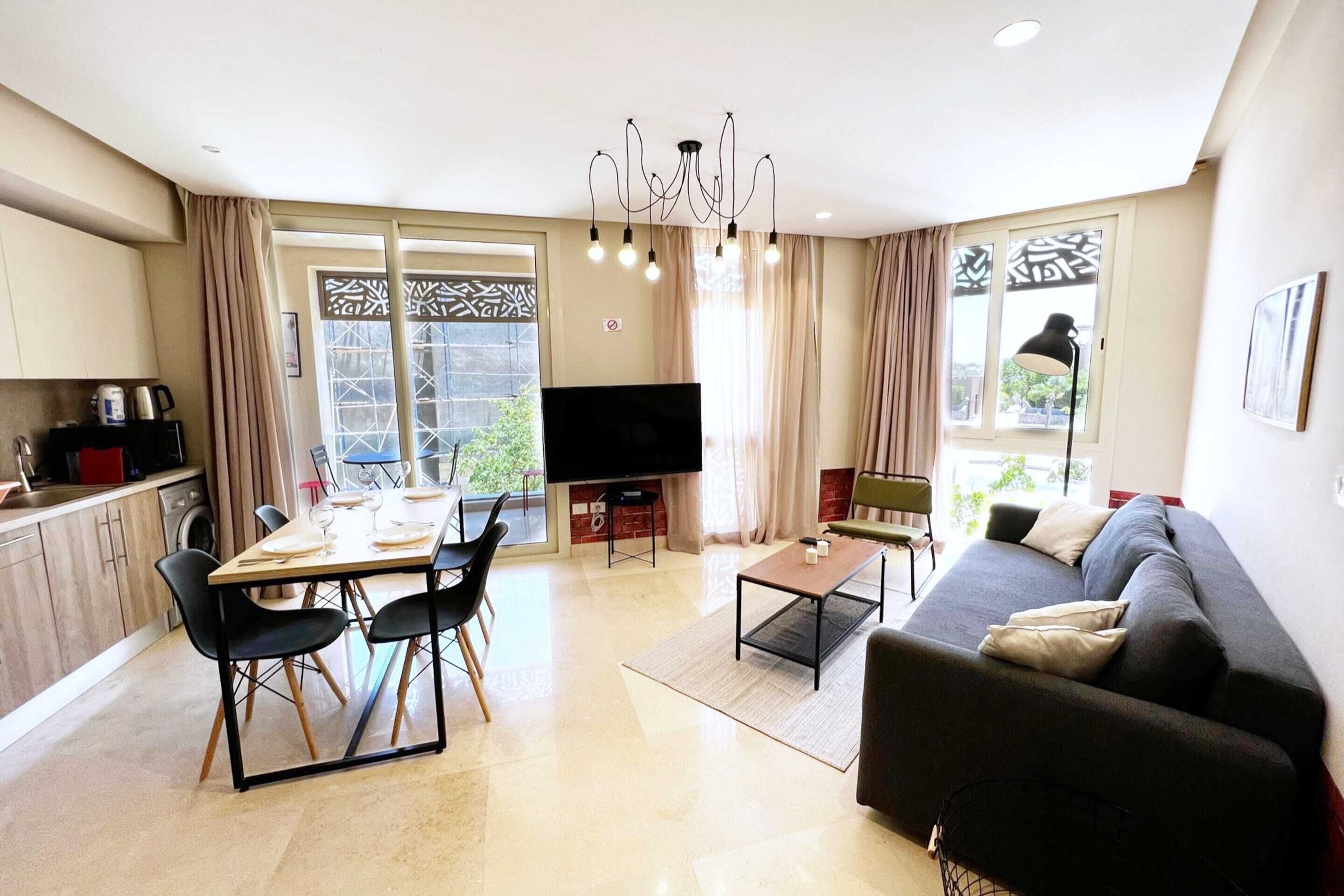 Een moderne woonkamer met een bank, een eettafel en stoelen. Er is een televisie, een kroonluchter en een raam met gordijnen.