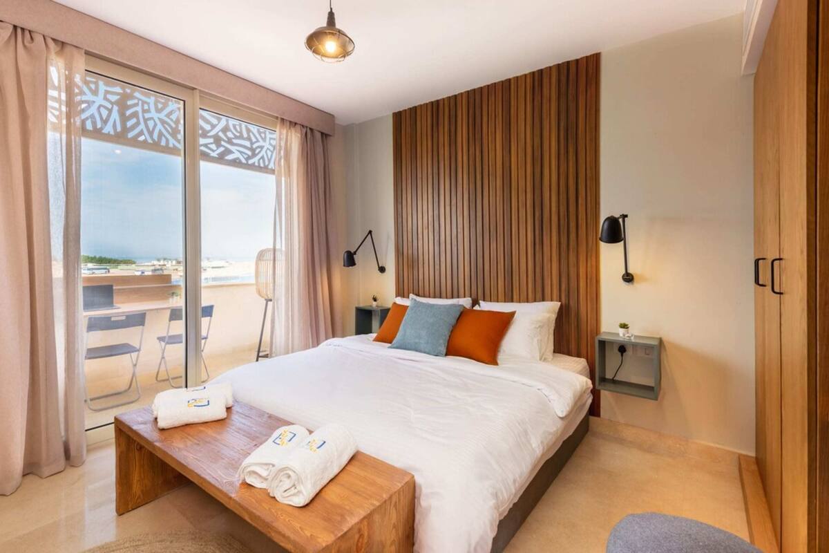 Une chambre d’hôtel avec un grand lit, une tête de lit en bois, un bureau avec une chaise, une table de chevet et une vue sur l’extérieur.