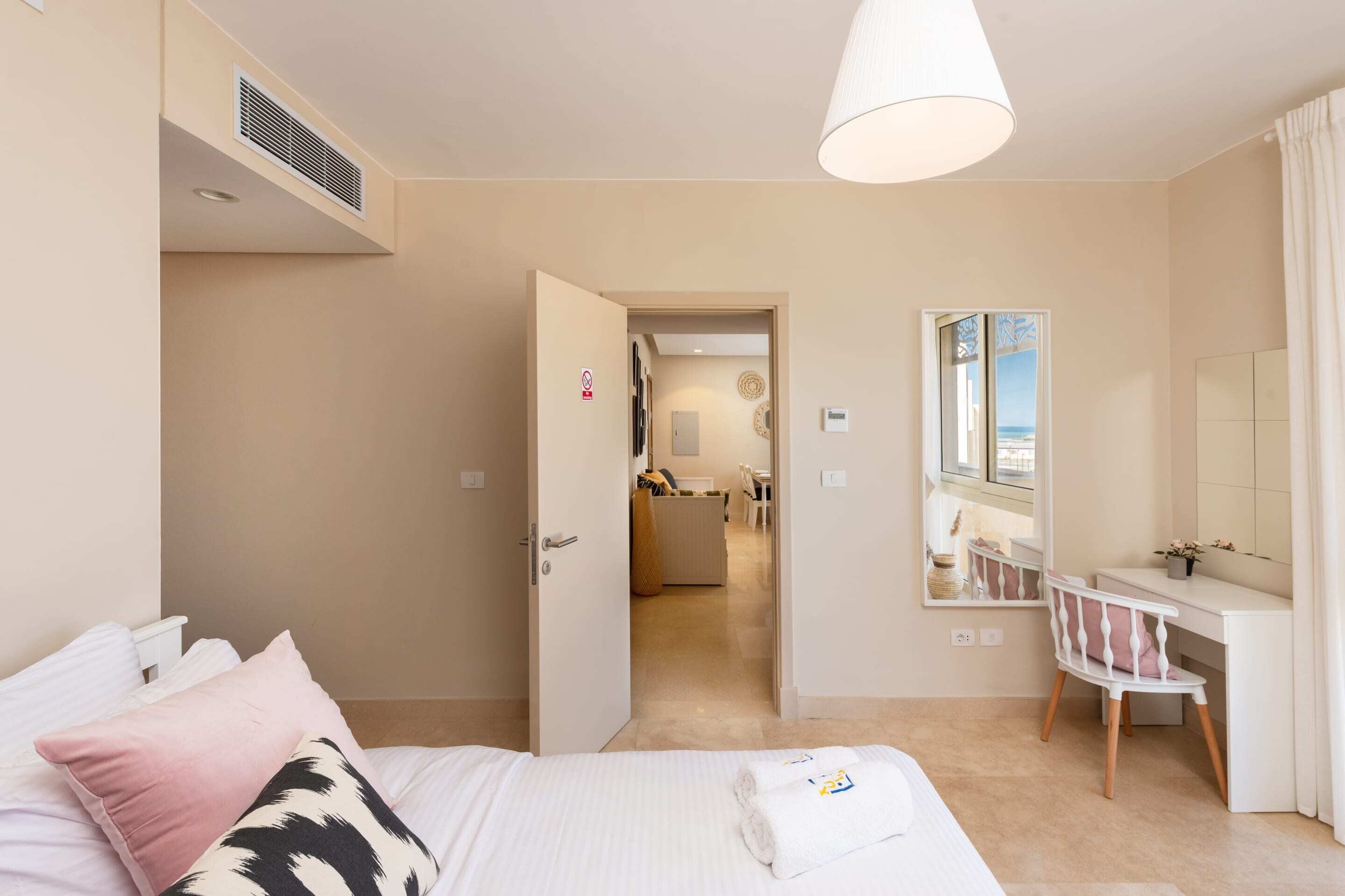 Ein Schlafzimmer mit einem Bett, einem Schreibtisch und einem Stuhl. Es gibt ein Badezimmer mit Spiegel und Waschbecken. Der Raum hat eine Tür zu einem anderen Zimmer.