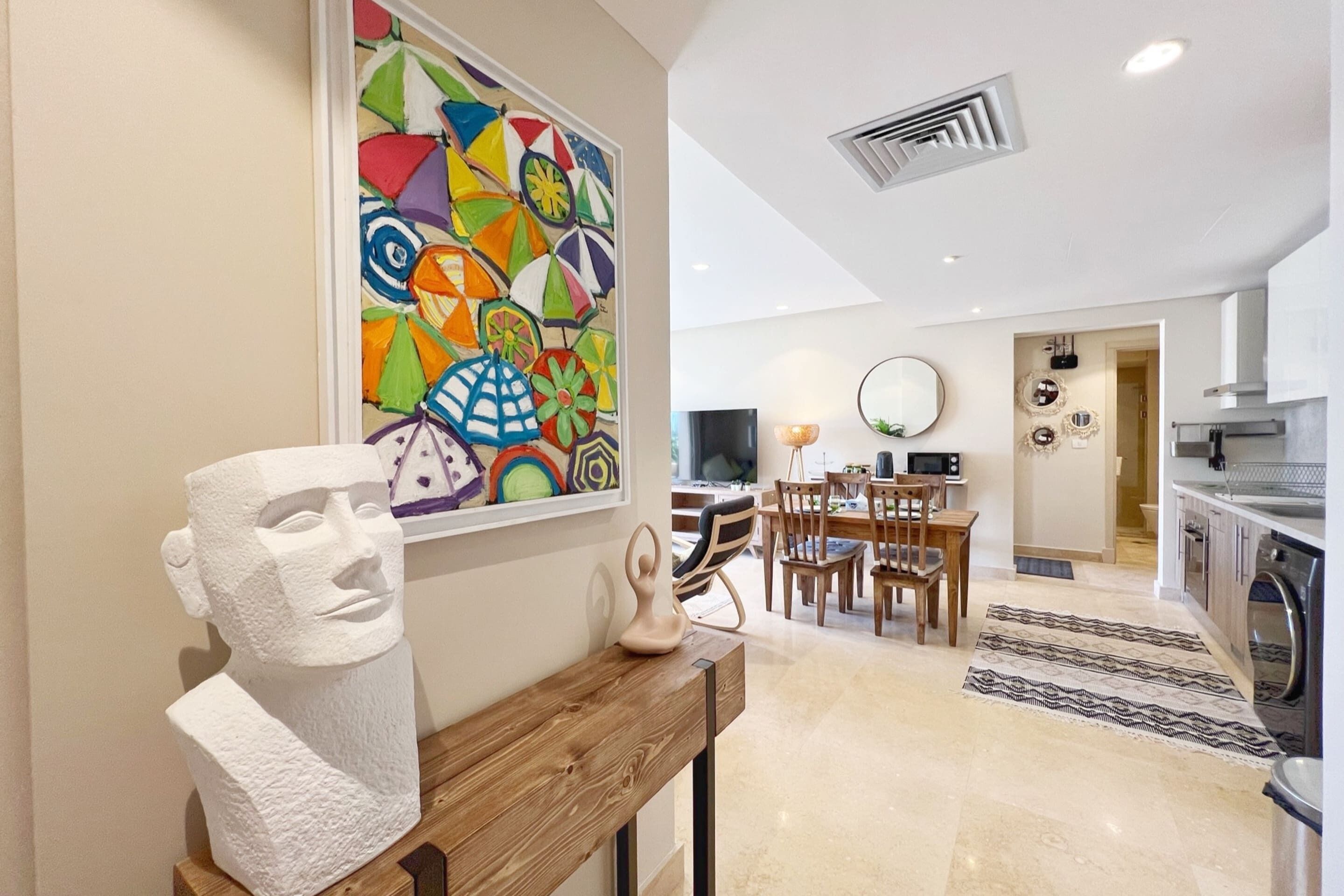 A modern kitchen with a dining area, a large colorful artwork on the wall, and a sculpture on a wooden table.
