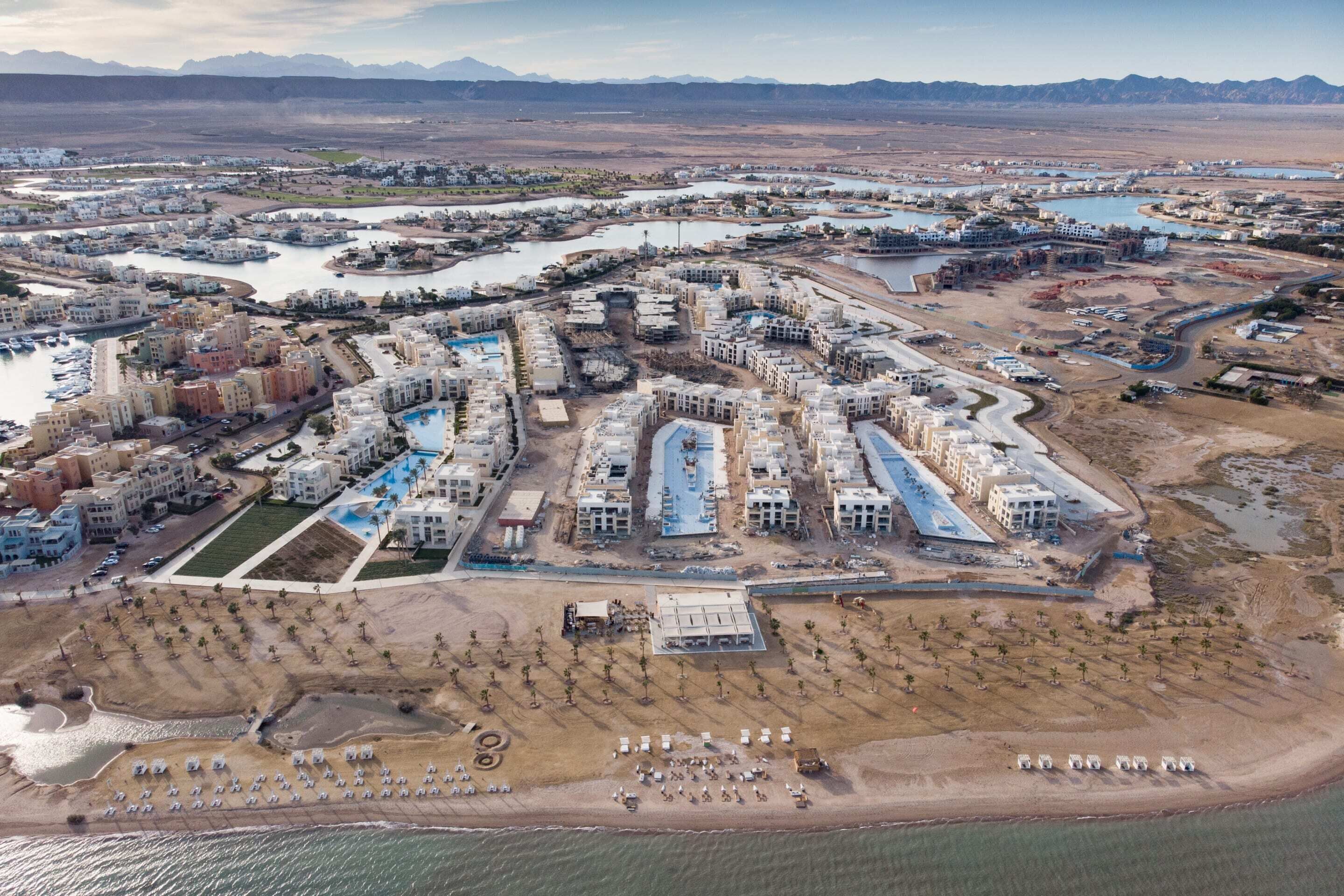Luftaufnahme eines Küstenresorts mit mehreren Gebäuden, einer Marina und einem Strand mit Liegestühlen.