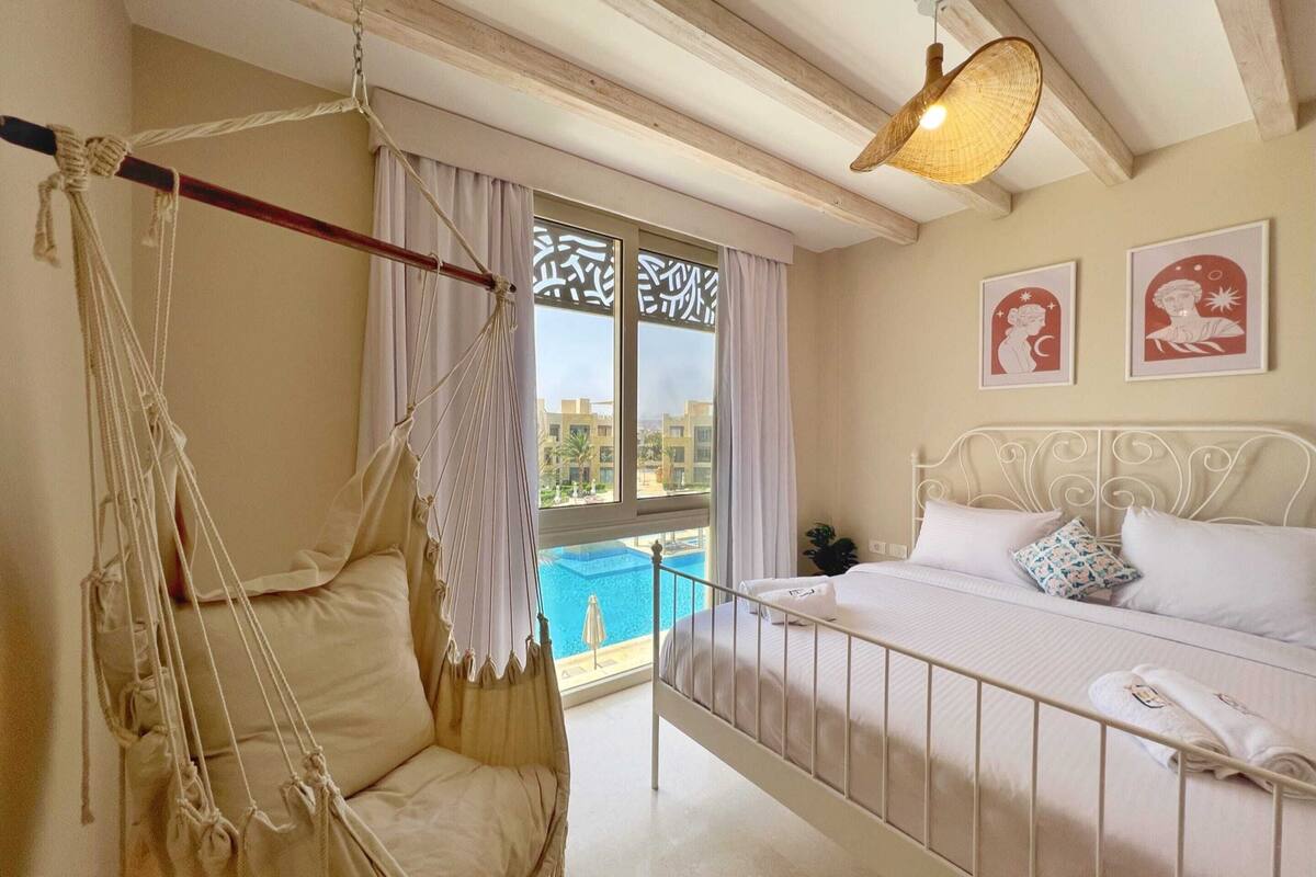 Une chambre avec un lit, un fauteuil suspendu et une vue sur une piscine.