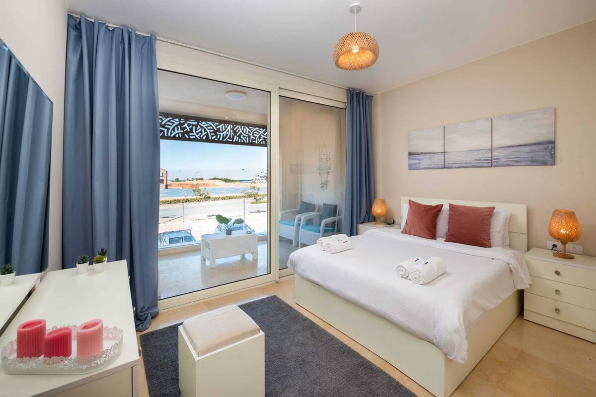 Une chambre d’hôtel moderne dotée d’un grand lit, d’un bureau, d’une chaise et offrant une vue sur la mer grâce à une porte coulissante.