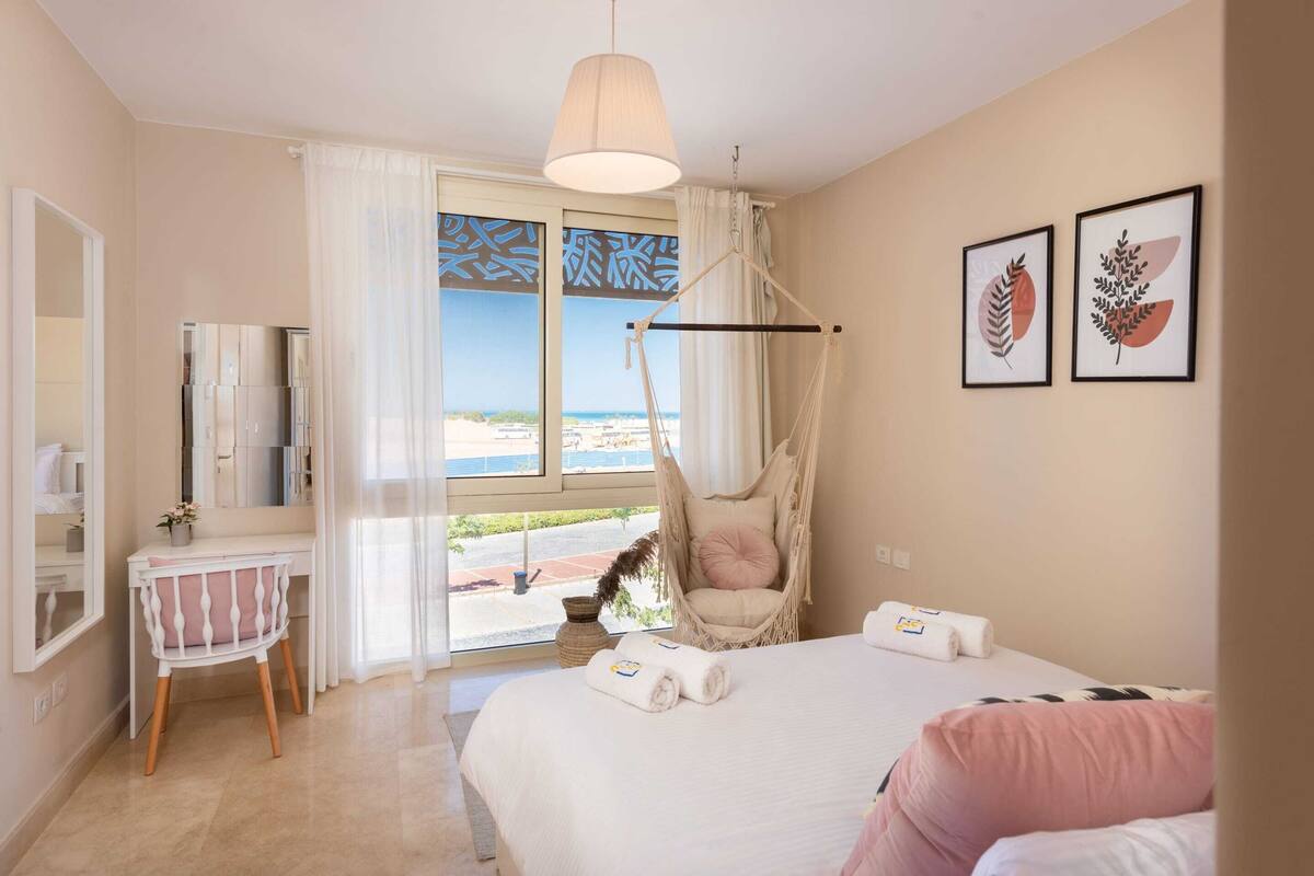 Une chambre avec un lit, une chaise, un bureau, un fauteuil suspendu et une vue sur la plage.