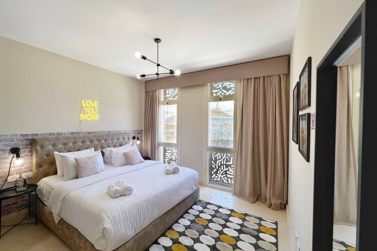Une chambre moderne avec une tête de lit capitonnée, un grand lit avec du linge de lit blanc, un néon mural et un tapis à motifs.