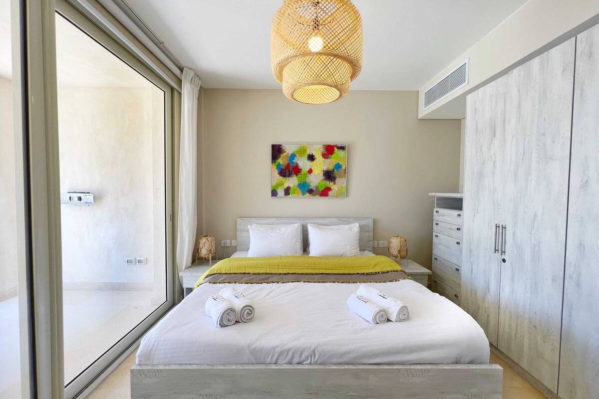 Une chambre à coucher moderne avec un grand lit, un luminaire suspendu en rotin, une peinture abstraite aux couleurs vives et une armoire.