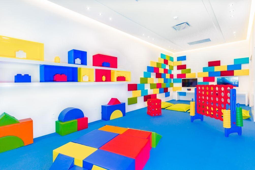 A brightly lit room with colorful geometric blocks on shelves and the floor, a television, and a red playpen.
