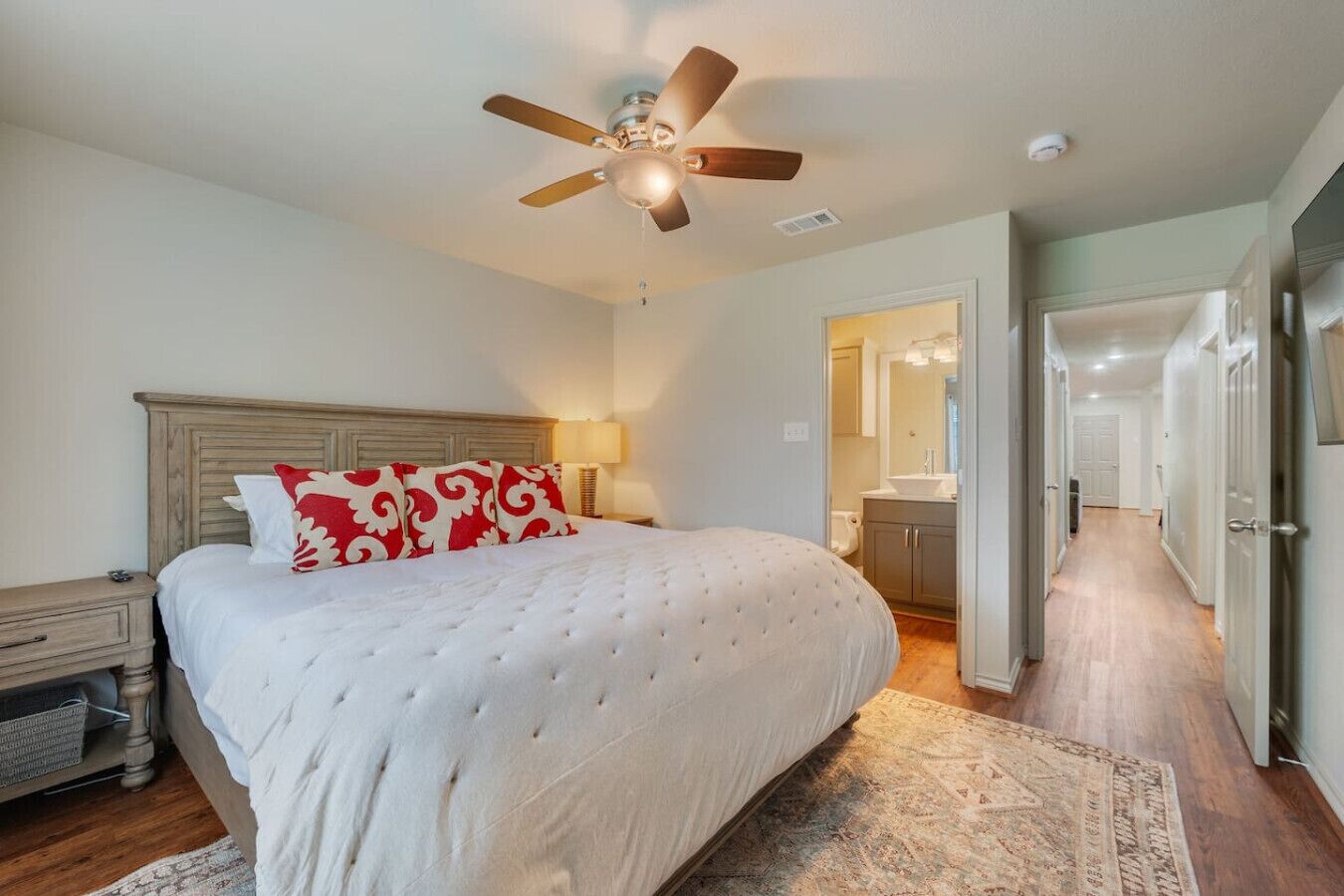 A bedroom with a large bed, a ceiling fan, a nightstand, a lamp, and a view into the bathroom.