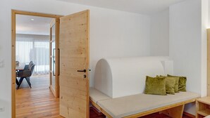 Een kamer met een houten deur, een witte muur, een bank met kussens en een eethoek op de achtergrond.