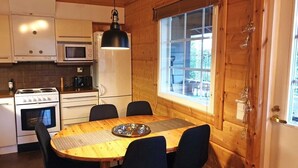 A wooden interior with a round dining table, black chairs, a black pendant light, a microwave, a stove, and a window with blinds. - Villa Slalom 2, Himos Länsihuippu (Keski-Suomi)