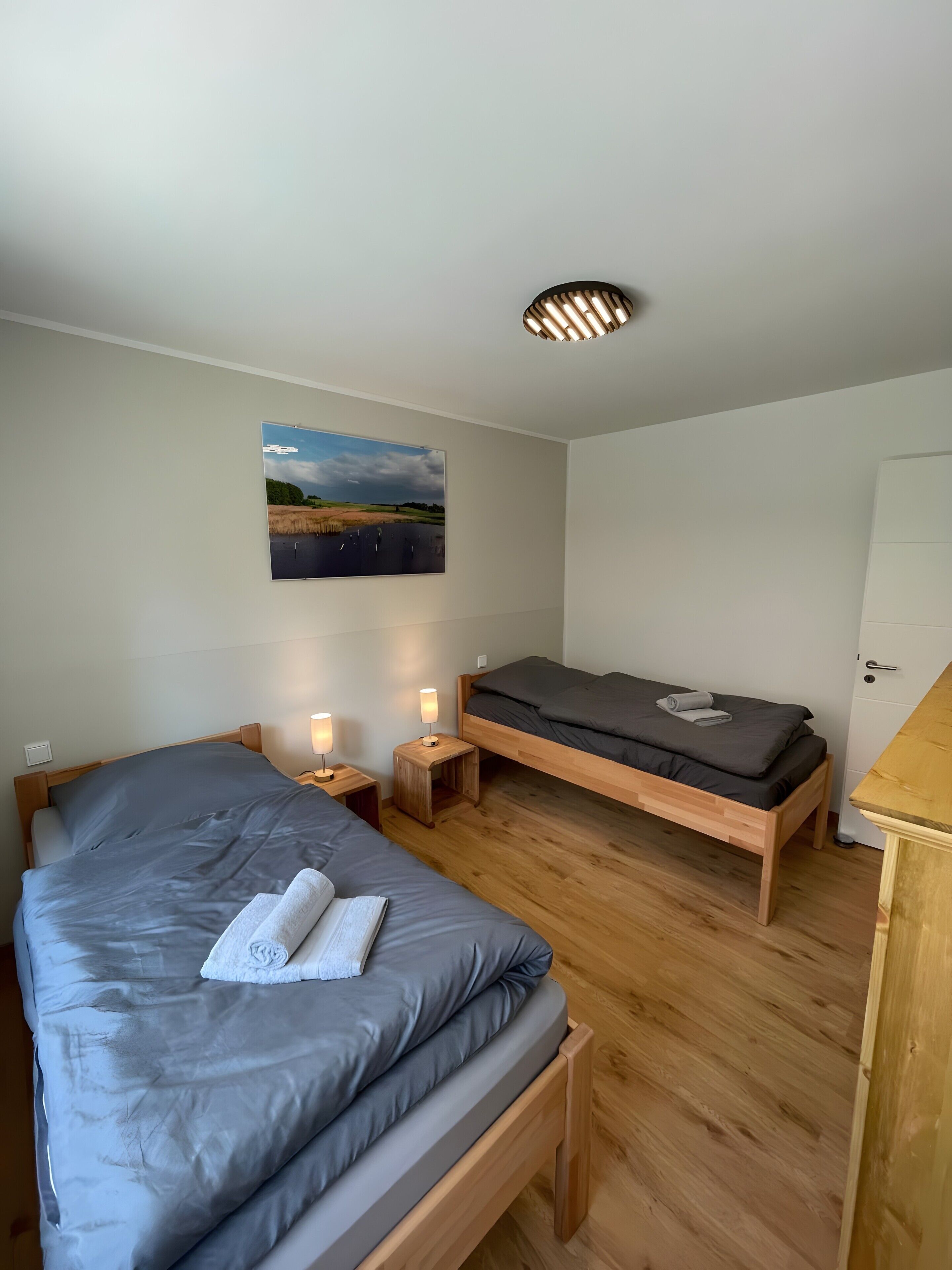 A bedroom with a large bed, a smaller bed, wooden flooring, bedside lamps, a painting on the wall, and a door.