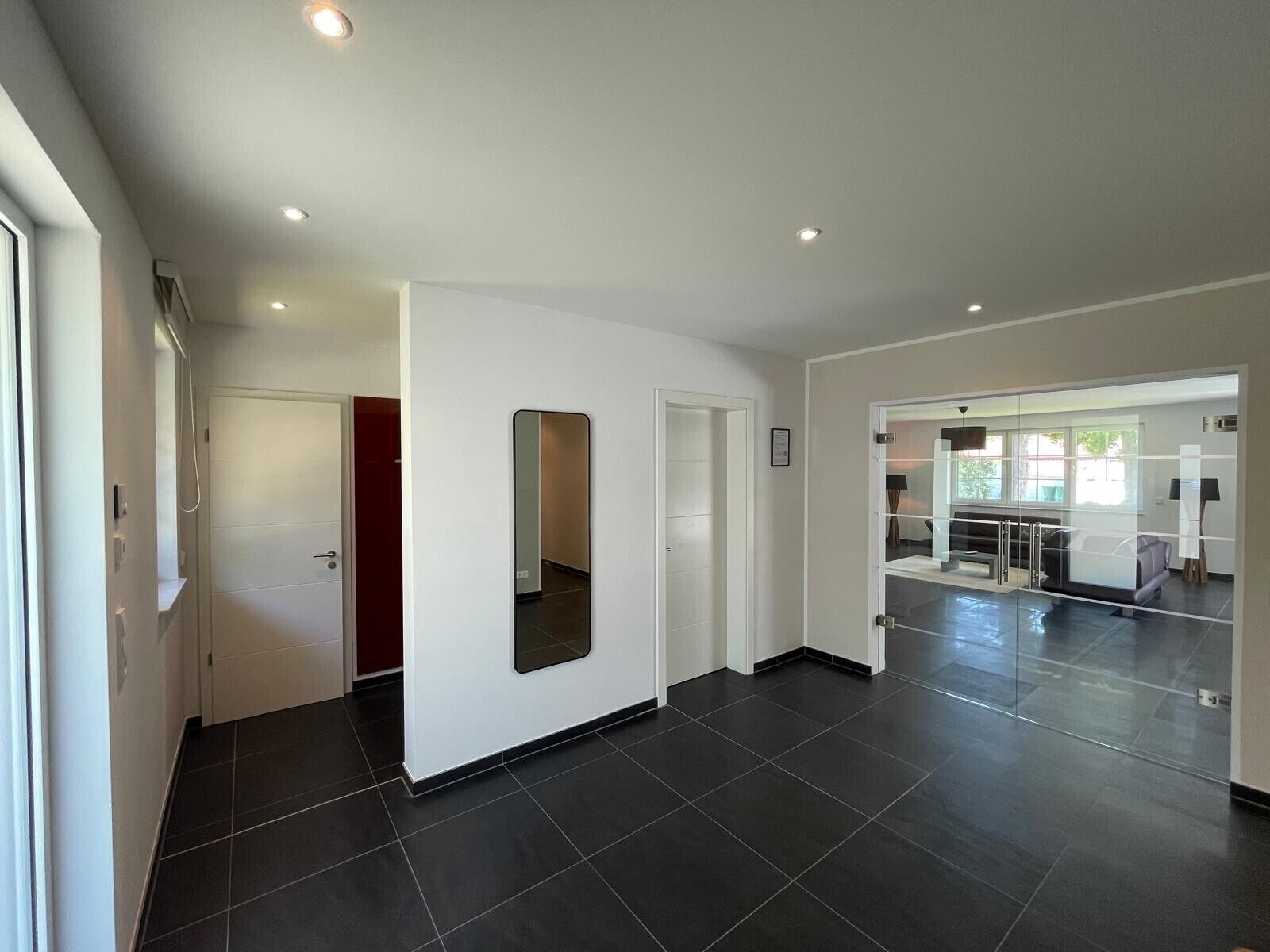A modern interior with a hallway leading to a living area, featuring a glass partition, recessed ceiling lights, and a tiled floor.