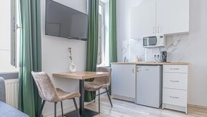 A compact living space with a kitchenette, a dining area with two chairs, a flat-screen TV mounted on the wall, and a window with green curtains. - Vienna Apts & Rooms in Prime Location (Vienna)