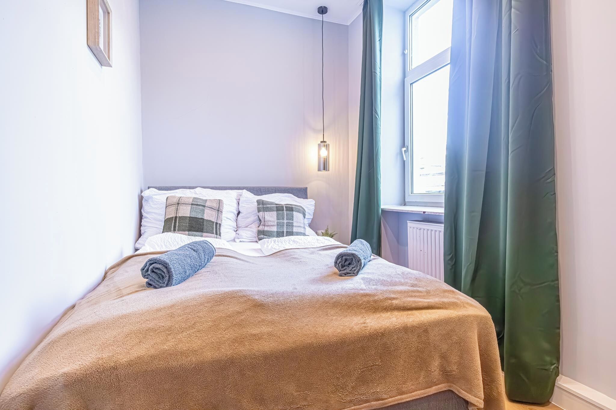 Una cama bien hecha con una manta marrón y dos toallas enrolladas, una lámpara de noche y una ventana con cortinas verdes.