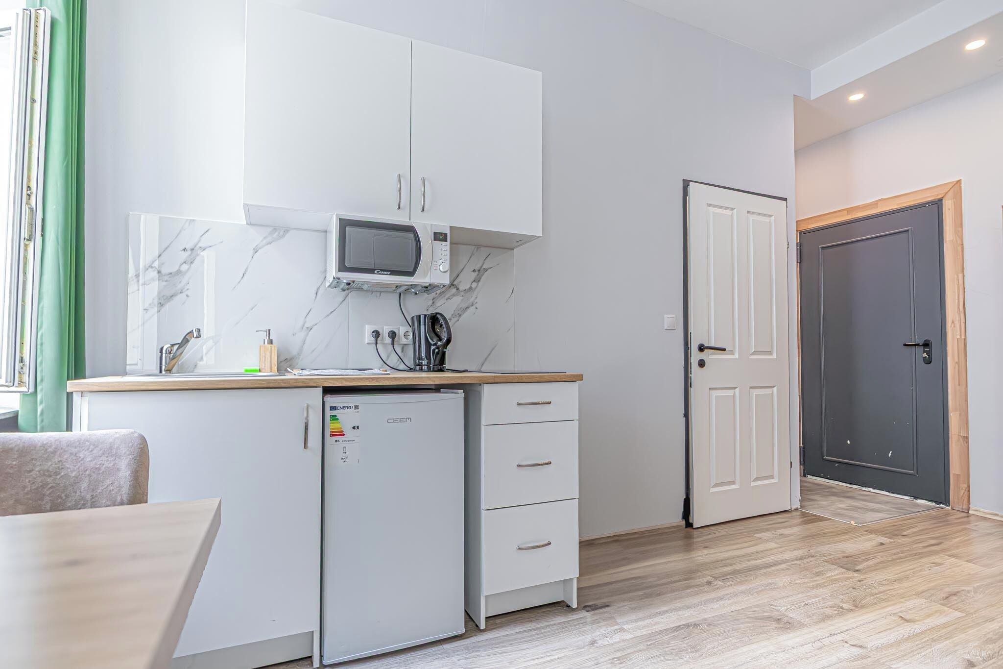 Eine kompakte Küche mit weißen Schränken, einer Mikrowelle, einem kleinen Kühlschrank und einer Holz Arbeitsplatte.