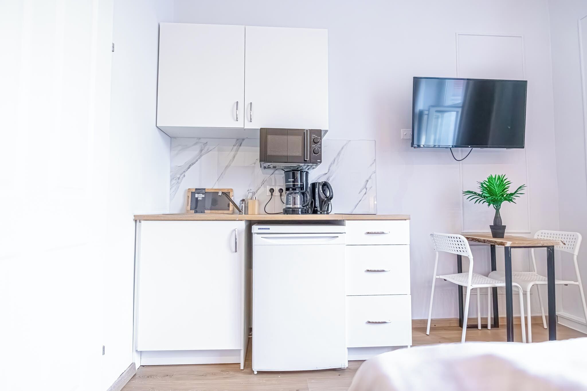 Cocina funcional con armarios blancos, microondas, cafetera y un refrigerador pequeño. Hay un comedor con mesa y sillas, y una planta en maceta en la pared.