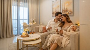 A couple sitting on a sofa in a well-lit room with a dining table and chairs, a chandelier, and artwork on the wall. - Keyrock Living - Oasis with Balcony View (Dubai)