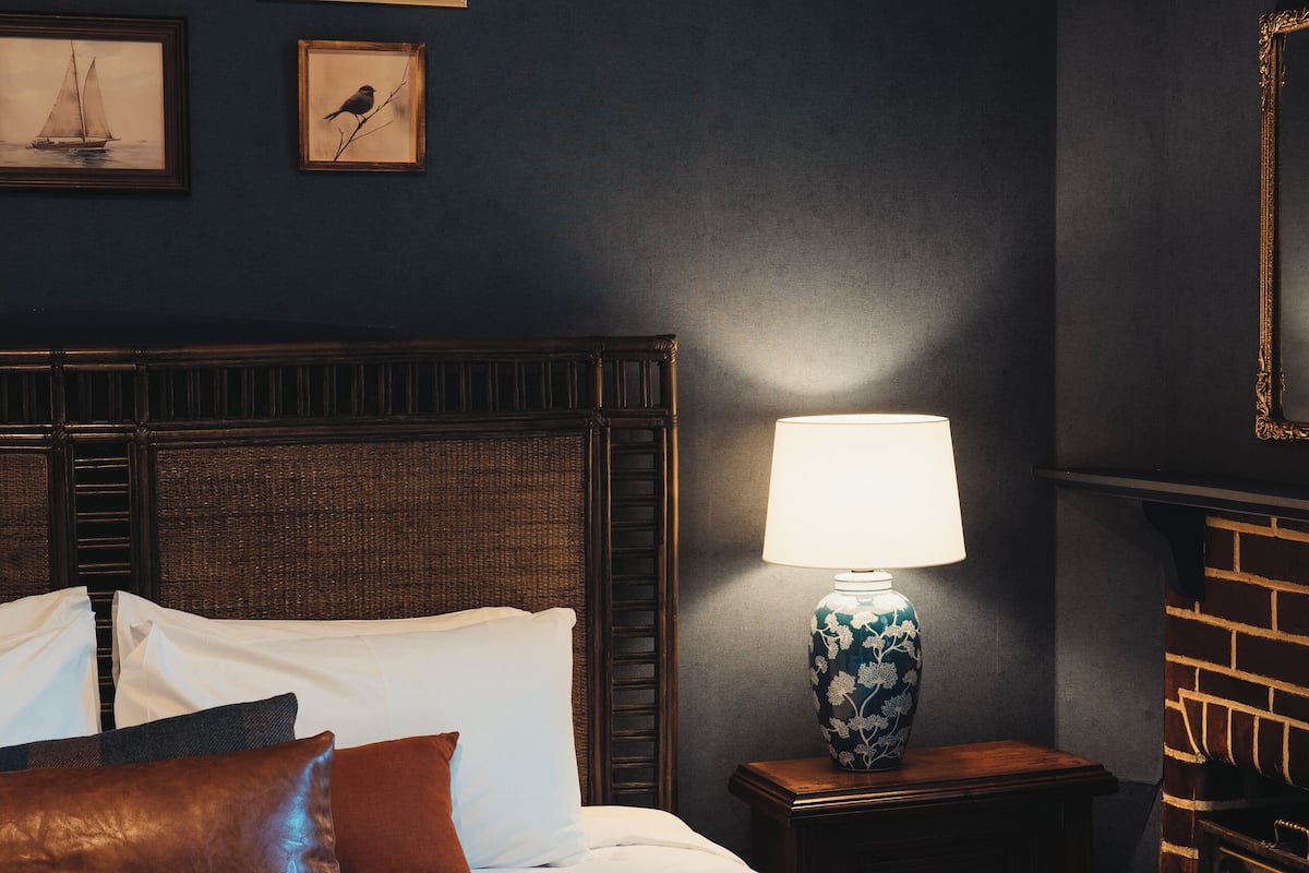 Ein ordentlich bezogenes Bett mit einer Karo-Decke, eine Lampe auf einem Nachttisch und gerahmte Bilder an der Wand.