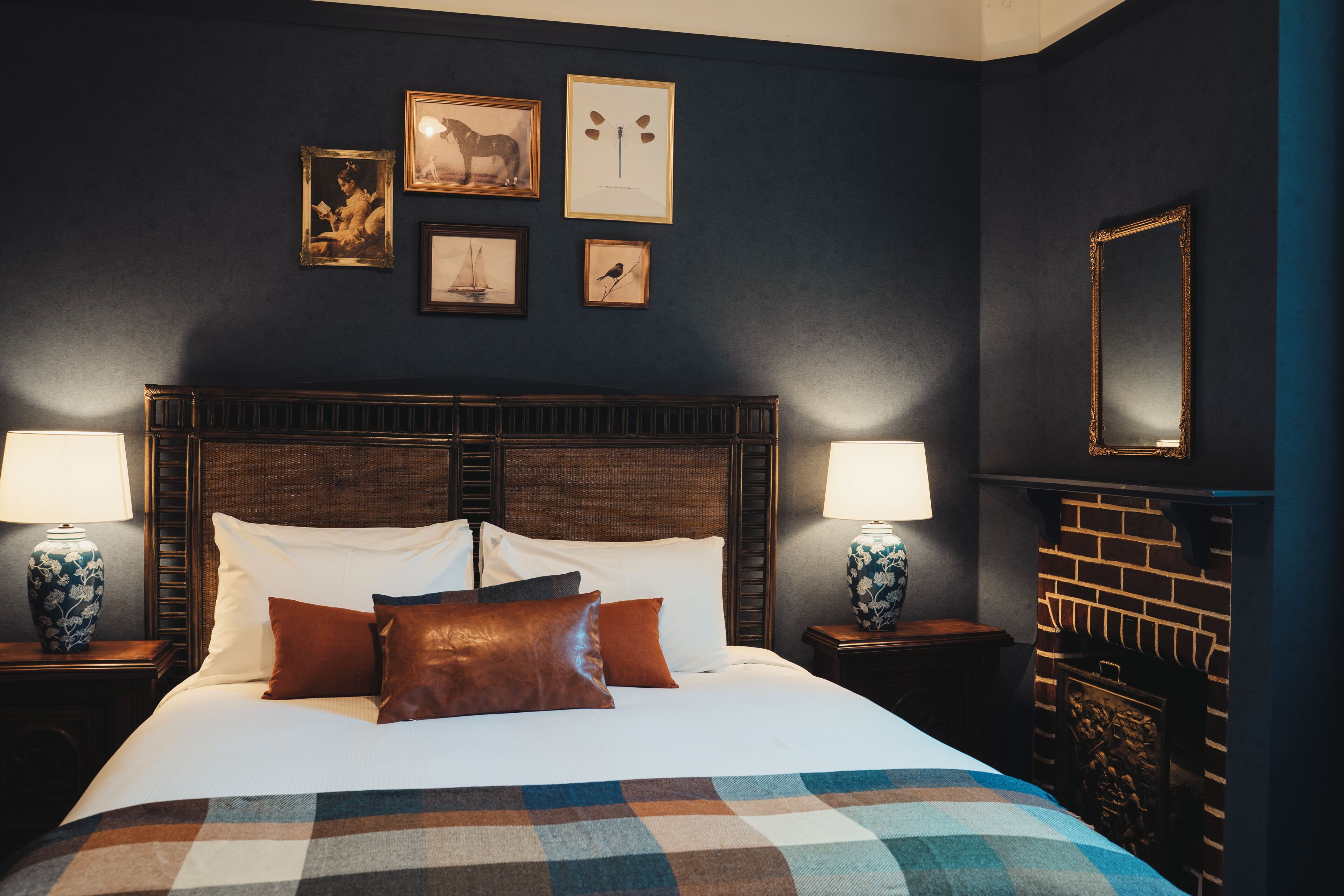 A neatly made bed with a checkered blanket, two bedside lamps, and a fireplace with a mirror.