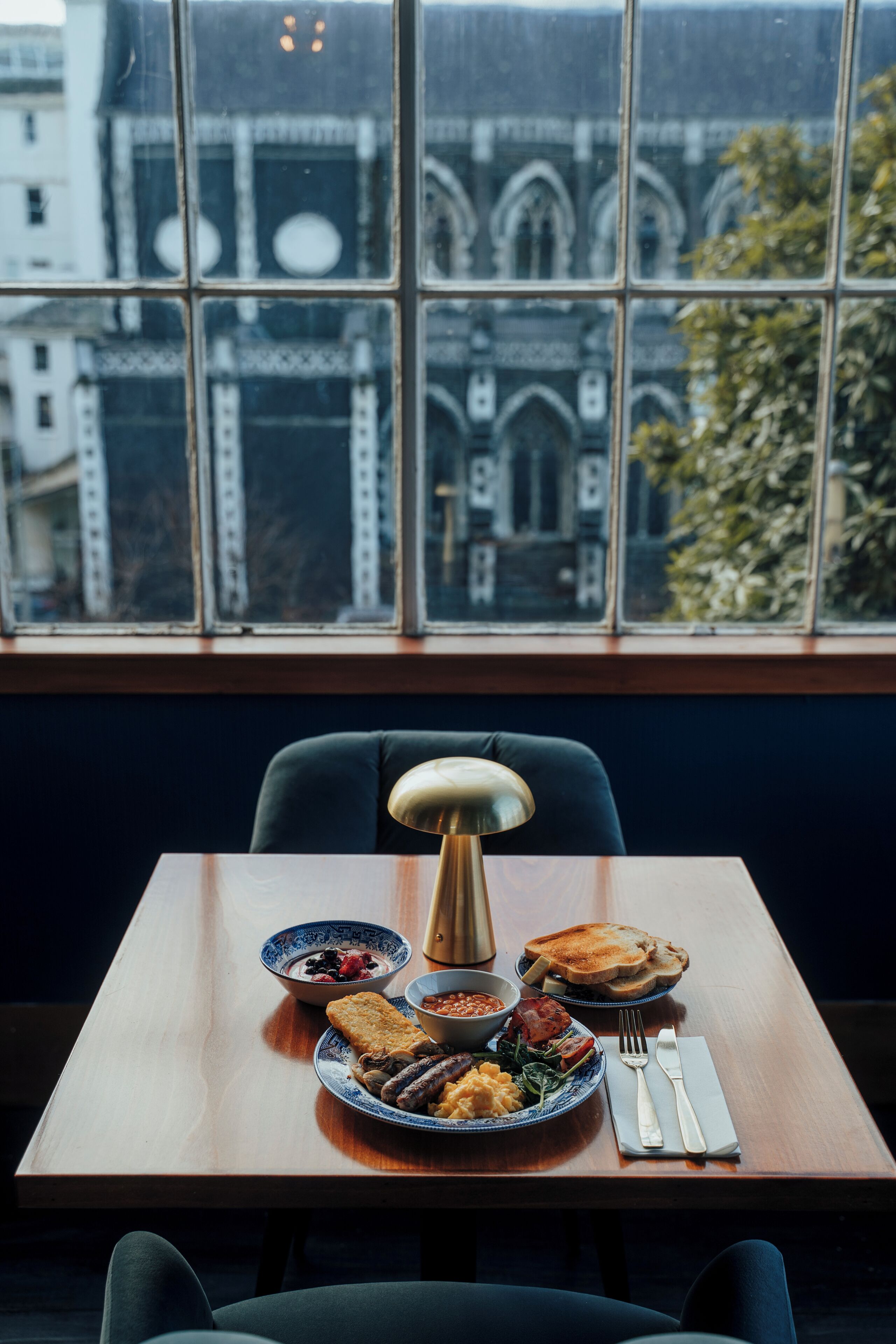 Une table à manger avec des assiettes remplies de nourriture, une lampe et des couverts. Une fenêtre donnant sur des immeubles se trouve à l’arrière-plan.