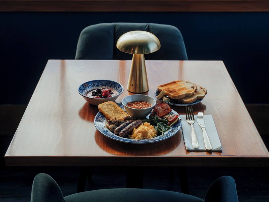 A dining table with plates of food, a lamp, and cutlery. A window with a view of buildings is in the background.