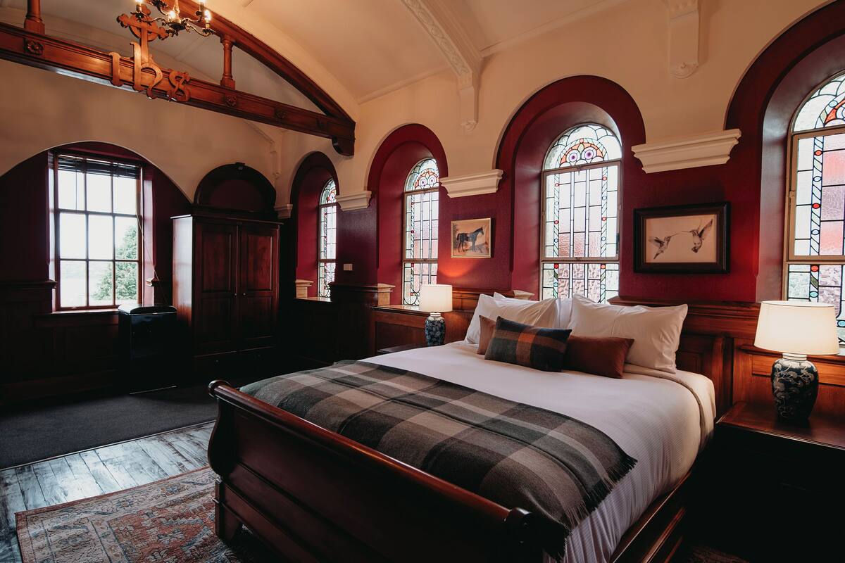 Ein Hotelzimmer mit einem großen Bett, einem Holz-Kopfteil und einer karierten Bettdecke. Zwei Nachttische mit Lampen, ein großes Fenster mit Buntglas und eine hölzerne Bank sind ebenfalls vorhanden.