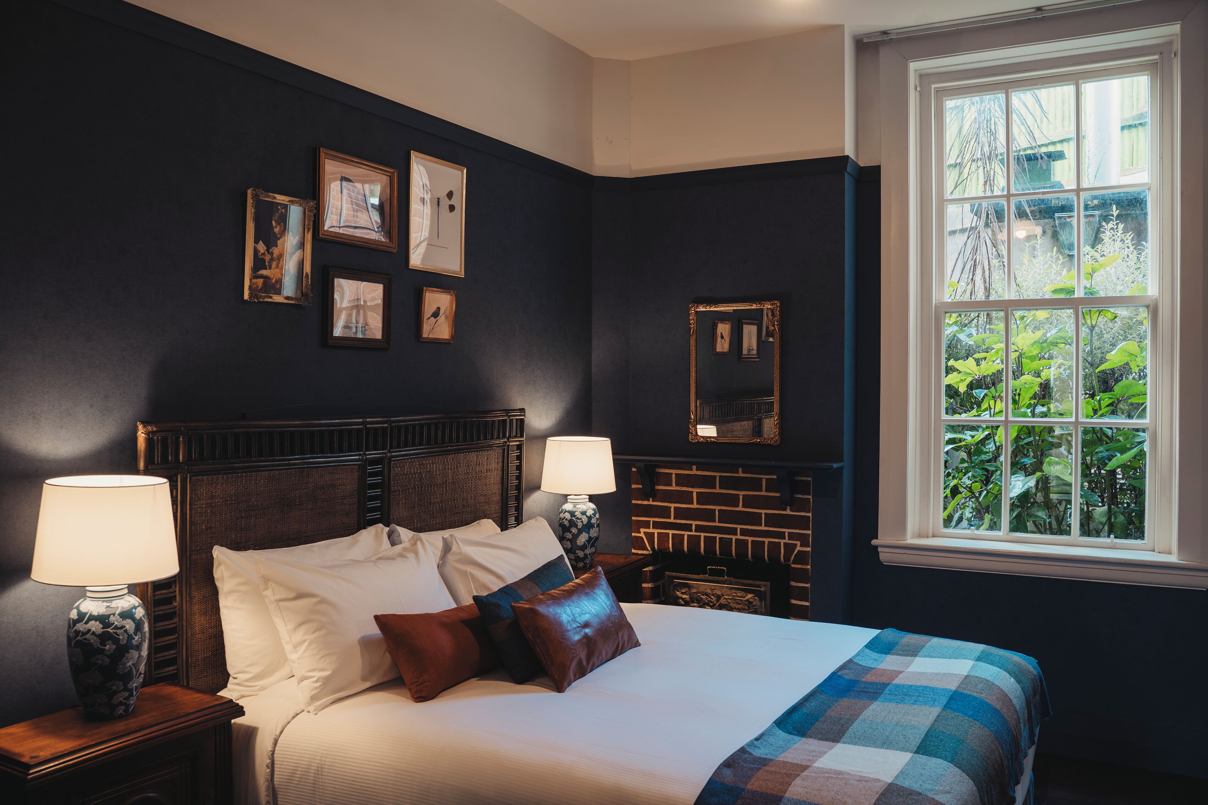 A bedroom with a large bed, two bedside tables, a nightstand, a brick fireplace, and a window with a view of greenery.