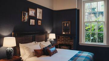 A bedroom with a large bed, two bedside tables, a nightstand, a brick fireplace, and a window with a view of greenery.