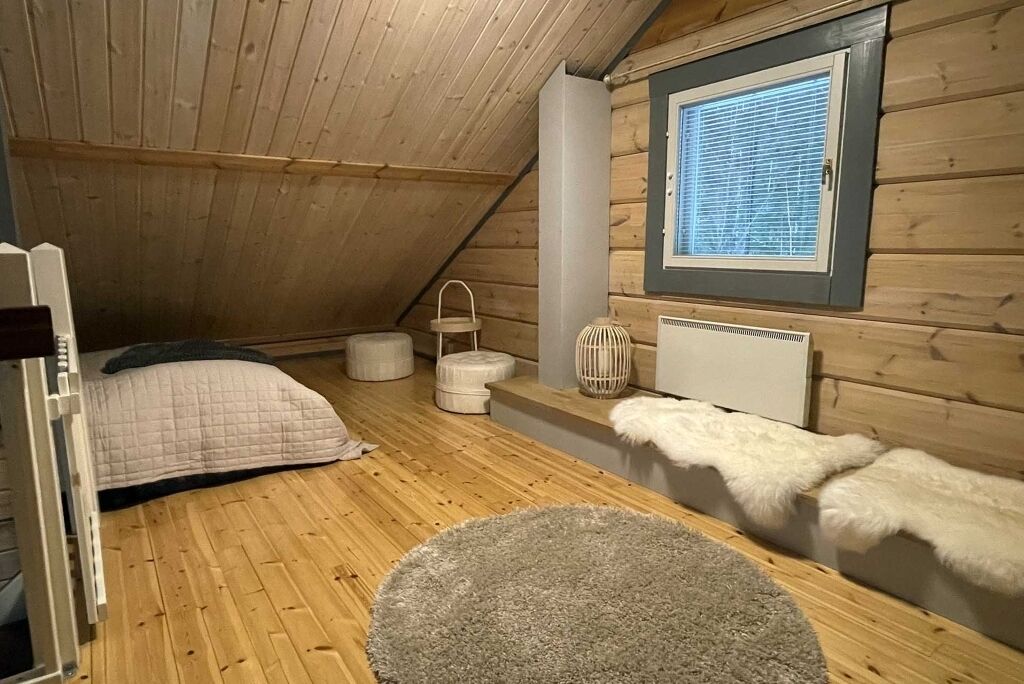 Une chambre mansardée chaleureuse, avec un plafond en pente, une grande fenêtre dotée de stores, un lit recouvert d’une couverture matelassée, un plaid blanc moelleux et un tapis rond.