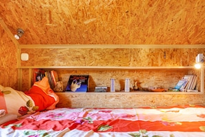 Een kamer met een houten plafond, een bed met een bloemenbedekking, een plank met boeken en decoratieve objecten, en een tafeltje met een kopje.