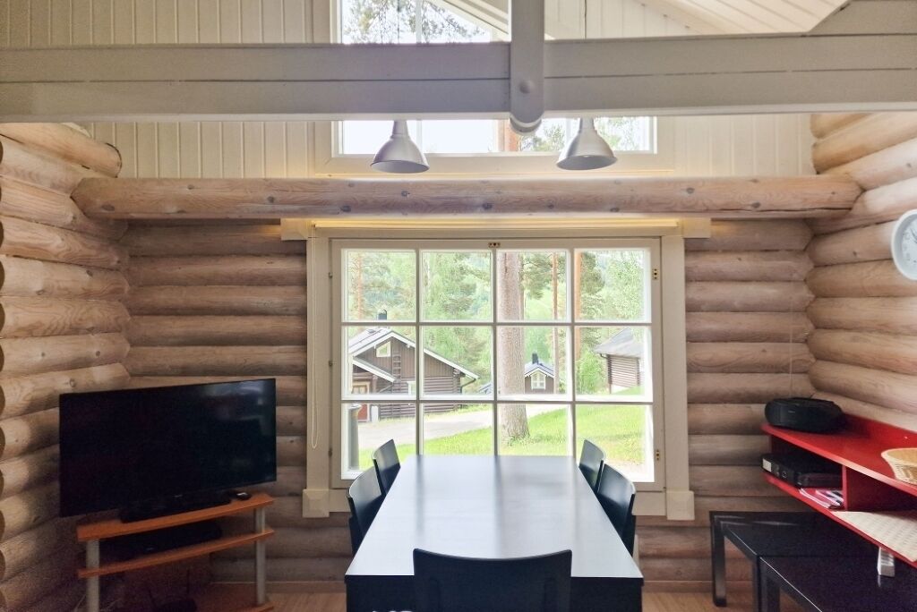 Habitación tipo cabaña de madera con un ventanal grande, una mesa de comedor, sillas, un televisor sobre un soporte de madera y un estante rojo con objetos.