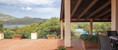 A covered terrace with a view of a lake and mountains, featuring potted plants and outdoor furniture.