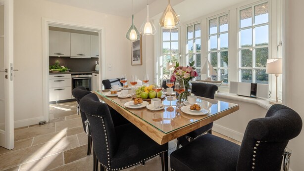 A dining area with a glass table, black upholstered chairs, and a view of the garden through large windows. - Finn's Hüs - A Stylish Thatched-Roof House in Wenningstedt on Sylt (Wenningstedt-Braderup (Sylt))