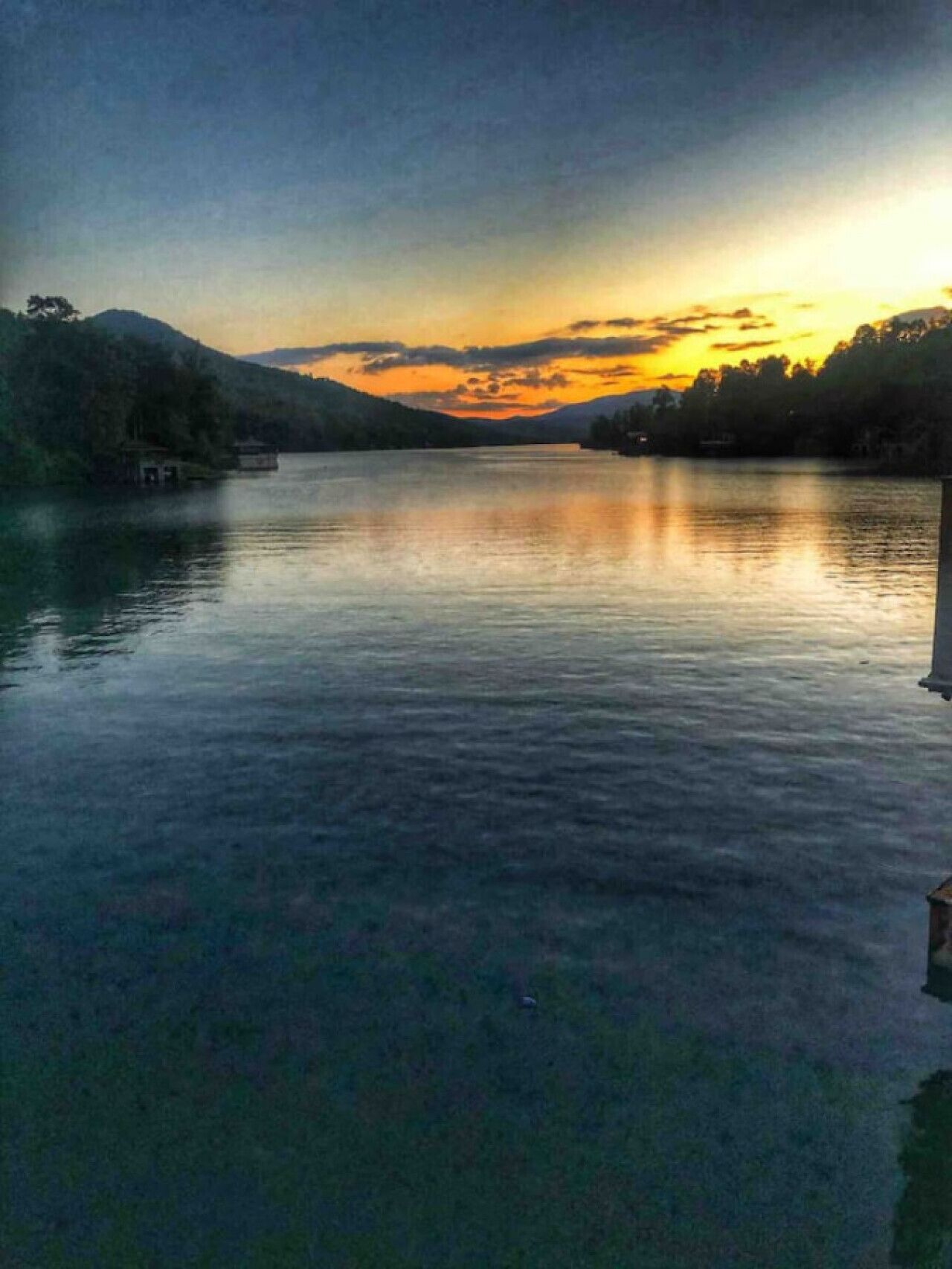 A calm lake with a wooden dock extending into the water, surrounded by hills and trees.